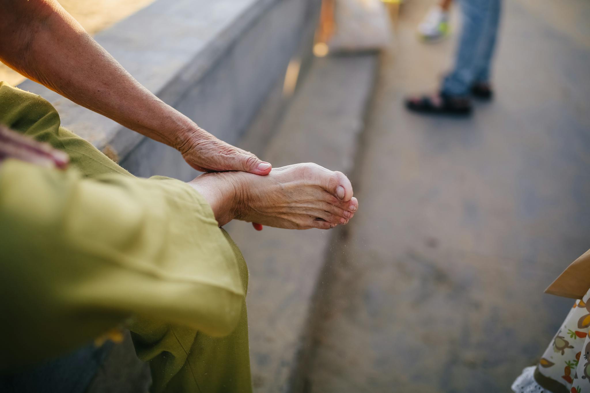 An elderly person sitting outdoors massaging their foot for relief.